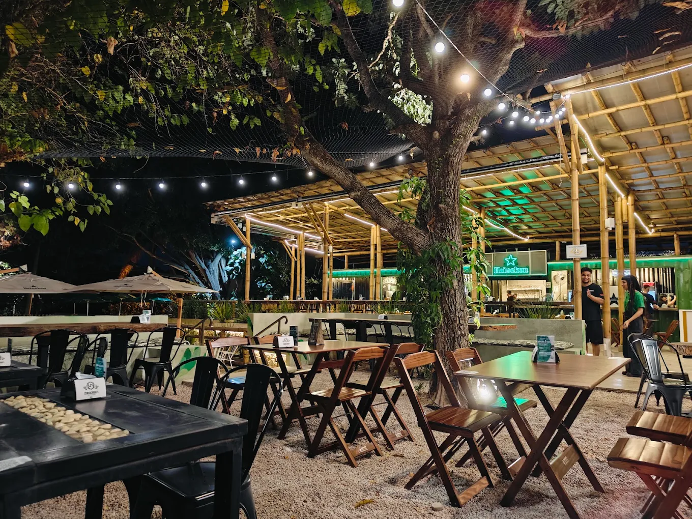 Quintal do Consagrado's à noite: árvore central iluminada por cordões de luz, cobertura de bambu e mesas de madeira sobre o cascalho