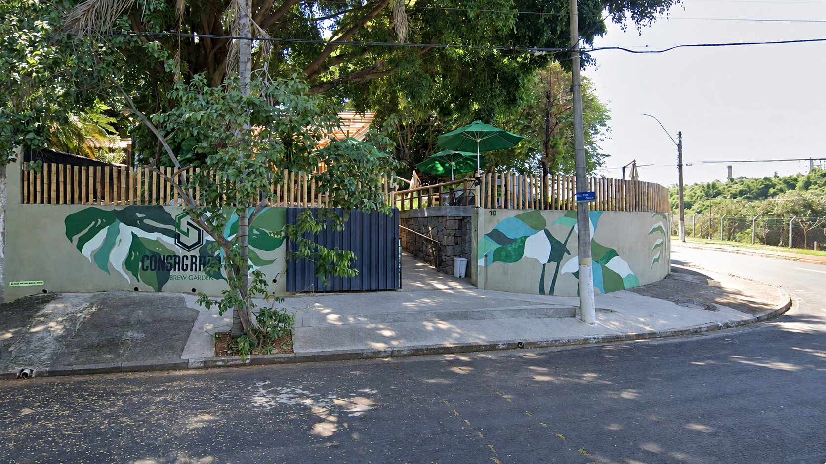 Entrada do Consagrado's Brew Garden na esquina da Rua Egas Muniz, com muro grafitado de folhagens e árvores sobre o quintal