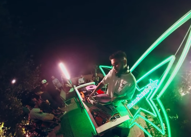 DJ no comando das pick-ups à noite no quintal, com a estrela de neon verde ao lado e o público em volta